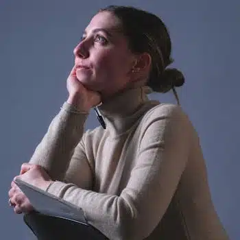 A thoughtful woman in a cozy sweater gazes upwards, holding a book, indoors.